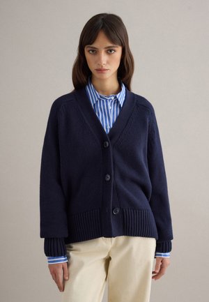 Donna che indossa un cardigan blu navy con bottoni sopra una camicia a righe blu e bianche e pantaloni beige, in piedi davanti a uno sfondo uniforme.
