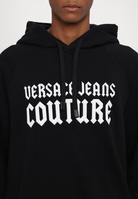 Μαύρη κουκούλα με λευκό κείμενο "VERSACE JEANS COUTURE" σε έντονη, γωνιακή γραμματοσειρά. Το υλικό είναι μαλακό βαμβάκι με λεία υφή.