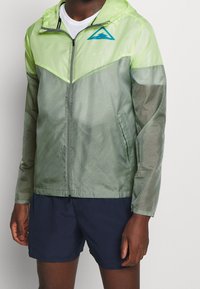 Lett, lynlåslukket jakke i gradient grøn med et trekantet logo; har hætte, tekstureret stof og sidelommer.