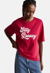 Czerwona bawełniana koszulka z napisem "Stay Groovy" w białym skrypcie. Luźny krój, krótkie rękawy, okrągły dekolt, połączona z niebieskimi dżinsami.