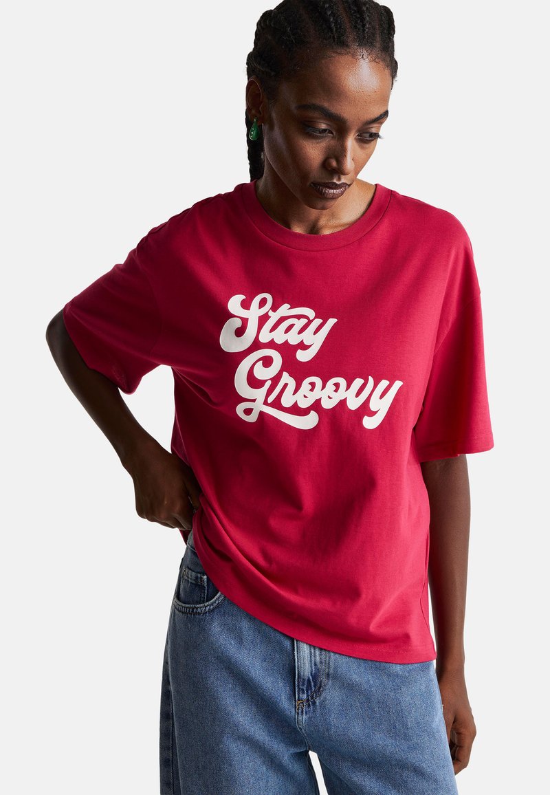 Czerwona bawełniana koszulka z napisem "Stay Groovy" w białym skrypcie. Luźny krój, krótkie rękawy, okrągły dekolt, połączona z niebieskimi dżinsami.
