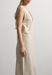 Beiges, ärmelloses Leinenkleid mit tiefem V-Ausschnitt und gerafftem Rückendetail, das eine sanfte, strukturierte Oberfläche und eine fließende Silhouette aufweist.