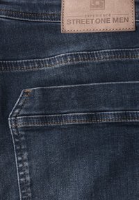 Un bolsillo trasero de un pantalón de mezclilla azul oscuro con costuras naranjas y una etiqueta de cuero marrón claro que dice "Street One Men."