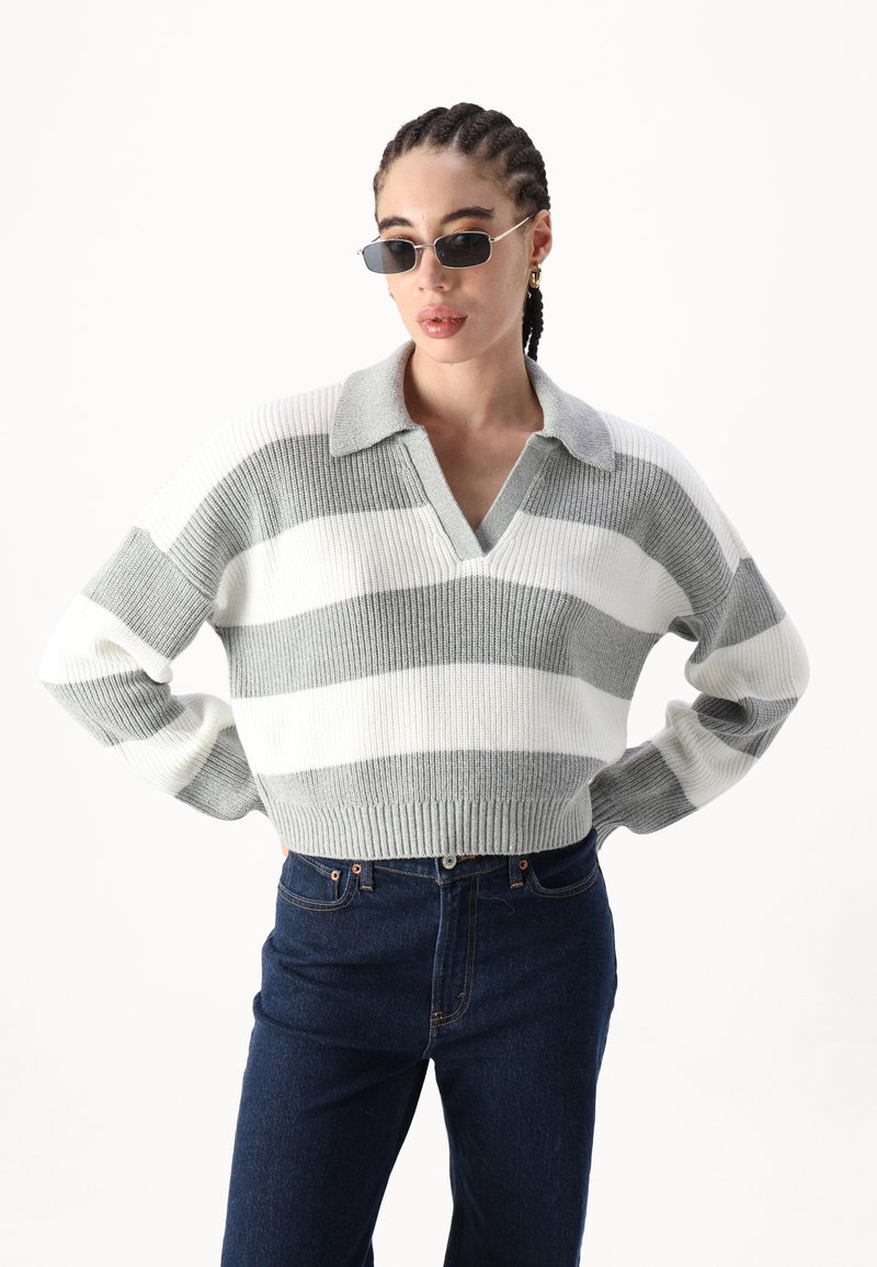 Szary i biały sweter w paski, dzianinowy, z dekoltem w kształcie litery V i kołnierzykiem, połączony z ciemnoniebieskimi dżinsami. Posiada ściągacz na dole i luźny krój.