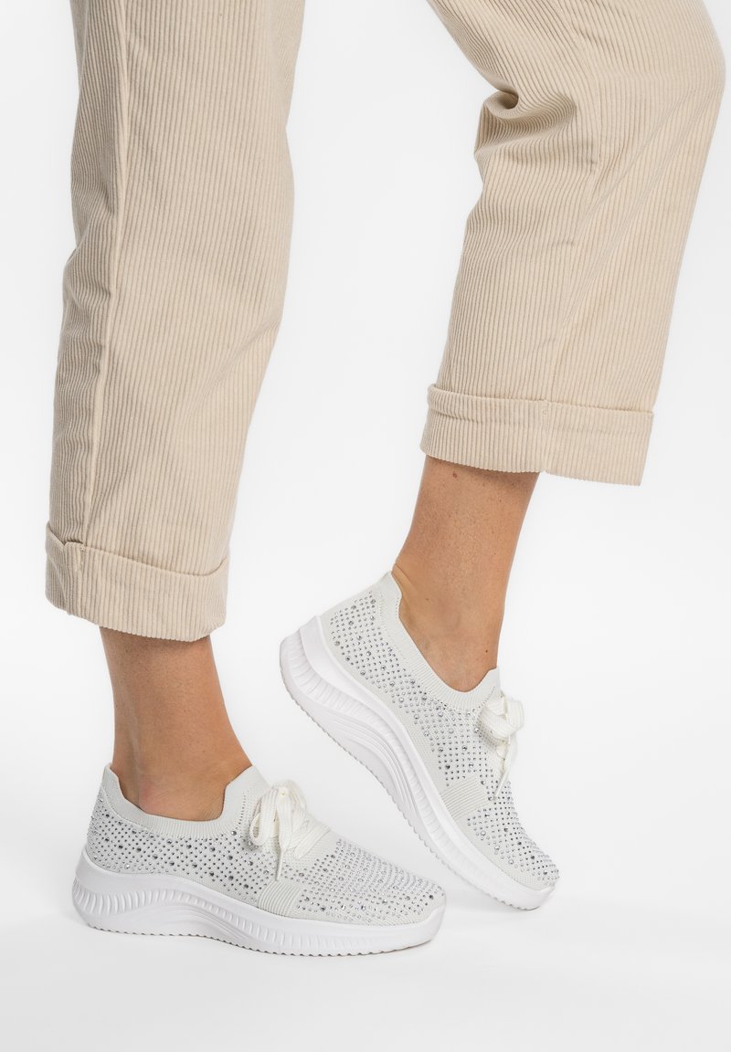 Gambe con pantaloni beige risvoltati e sneaker bianche lavorate a maglia con lacci e suola strutturata, posate su sfondo bianco.