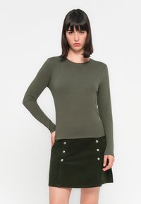 Haut à manches longues, de couleur vert olive, avec un col rond, associé à une jupe en velours côtelé verte avec des accents de boutons en argent et une poche latérale.