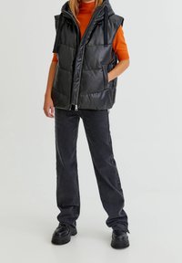 Doudoune noire sans manches avec capuche, munie de panneaux matelassés et de poches latérales. Portée sur un haut orange, associée à un jean gris foncé et des chaussures noires.