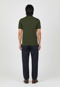Camiseta de polo de manga corta en verde oliva con textura suave, combinada con pantalones azul marino y zapatos marrones, vista desde la parte de atrás.