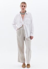 Camisa blanca de texture con botones y bolsillo delantero, combinada con pantalones de lino beige claro. Lleva zapatos de cuero trenzado color marrón claro.