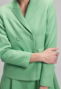 Žena v dvouřadém blazeru světle zelené barvy a odpovídajících kalhotách, s jednou rukou překříženou přes druhou na neutrálním pozadí.