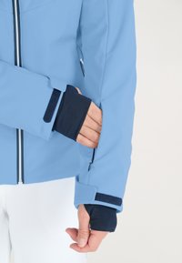 Lysblå vandafvisende jakke med m�ørkeblå inderside ved manchetterne, der har lynlås og velcro-lukning. Søm detaljer og sidelomme synlige.
