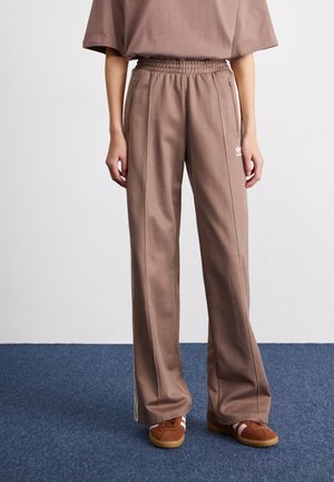 Personne portant un pantalon de survêtement large marron Adidas avec une taille élastique et des poches zippées, associé à des chaussures décontractées marron sur un tapis bleu.