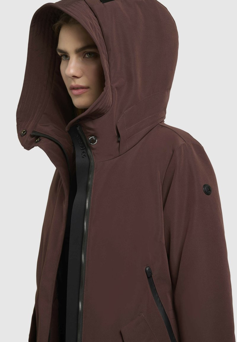 Giacca impermeabile marrone con un ampio cappuccio, chiusura con zip, tasche laterali e un accento nero sulla zip. Texture liscia e opaca in tutta la superficie.