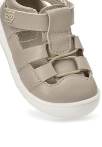 Sandalia de cuero beige con cordones elásticos, correas laterales y una parte superior acolchada. Presenta una suela de goma blanca y una etiqueta de marca en el lateral.