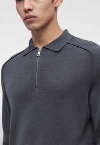 Jeune homme portant une chemise à manches longues gris foncé avec un petit col et une fermeture éclair argentée partiellement fermée à l'avant.