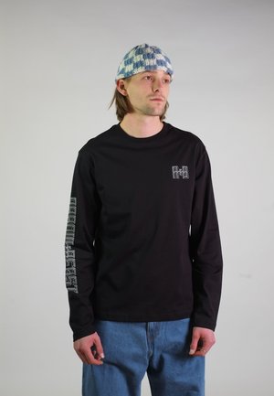 Junger Mann trägt eine blau-weiß karierte Strickmütze, ein schwarzes Langarmshirt mit weißem Text am Ärmel und blaue Jeans, steht vor einem grauen Hintergrund.