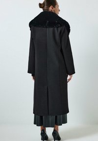 Vrouw in een lange zwarte jas met een grote bontkraag, zwarte midi-rok en zwarte hoge hakken, staande met haar rug naar de camera tegen een effen achtergrond.