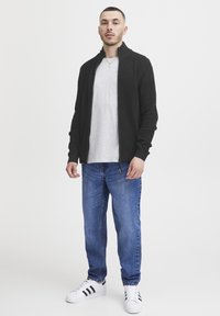 Svart teksturert zip-up genser over en grå t-skjorte, kombinert med blå jeans og hvite joggesko med svarte striper. Enkel, moderne design.
