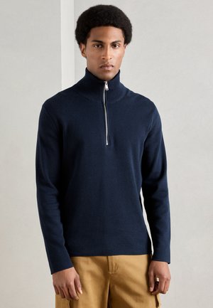 Man med kort lockigt hår som bär en marinblå zip-up polotröja och beige cargobyxor, stående mot en slät vägg.