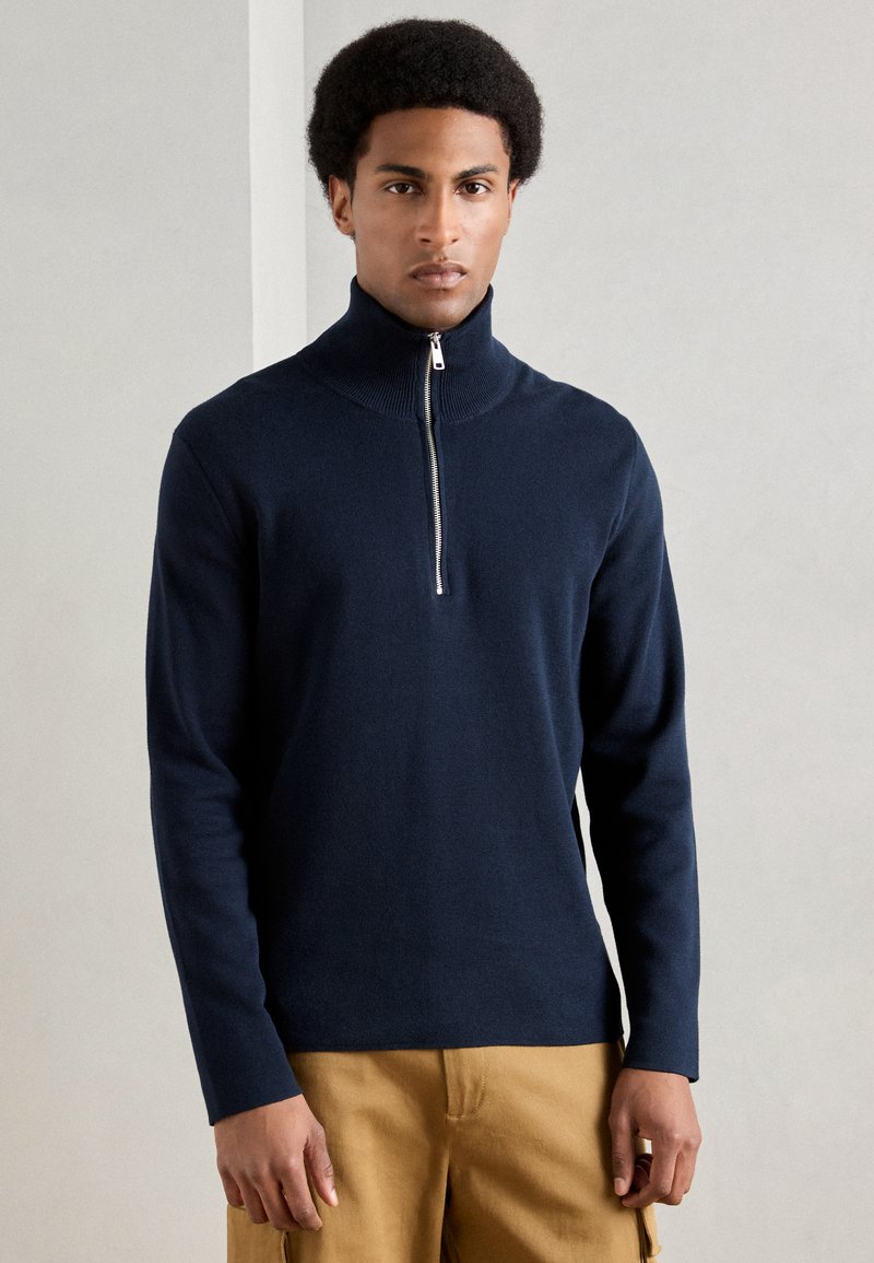 Άνδρας με κοντά σγουρά μαλλιά, φορώντας σκούρο μπλε φούτερ με κολάρο που κλείνει με φερμουάρ και μπεζ cargo παντελόνι, στέκεται μπροστά σε μια απλή τοίχο.