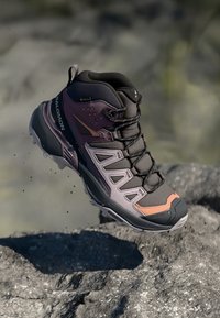 Botă de drumeție cu un upper sintetic gri și violet, accente portocalii, talpă din cauciuc texturată și sistem de șireturi. Caracteristici de design impermeabil.