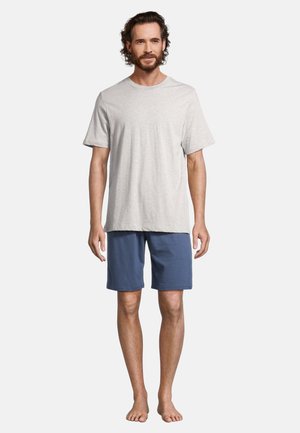 Mand med skæg iført en lysegrå kortærmet T-shirt og blå knælange shorts, stående barfodet mod en ensfarvet baggrund.