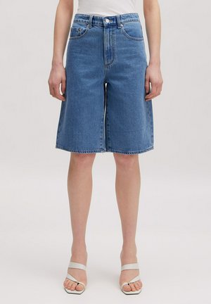 Frau trägt mittellange blaue Jeansshorts und weiße Riemchensandaletten mit hohen Absätzen, steht vor einem schlichten Hintergrund.