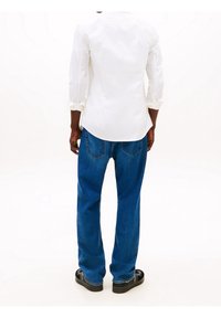 Camisa blanca abotonada con las mangas remangadas, combinada con vaqueros azules sueltos y sandalias negras, vista desde atrás.