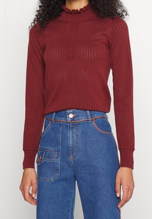 Nainen, jolla on päällään istuva viininpunainen neulepaita, jossa on teksturoitu kuvio ja röyhelökaulus, yhdistettynä korkeavyötäröisiin sinisiin farkkuihin, joissa on sivutasku.