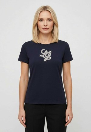 Donna bionda che indossa una t-shirt blu navy con lettere scintillanti "LO" sul petto e pantaloni neri, in piedi su uno sfondo chiaro.