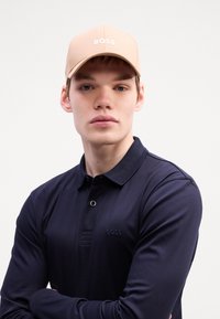 Beige Kappe mit "BOSS"-Logo, marineblaues Langarm-Polohemd mit subtiler Struktur und Knopfdetail, Model posiert mit verschränkten Armen.