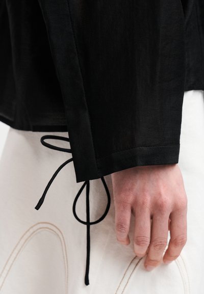 Veste noire et légère à la texture lisse. Comprend un détail de lien au niveau du poignet. Portée par-dessus une jupe texturée de couleur claire.