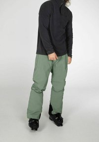 Maglione in pile nero a maniche lunghe con zip metà, abbinato a pantaloni da neve verde chiaro e scarponi da sci neri. Tessuto testurizzato, design funzionale.