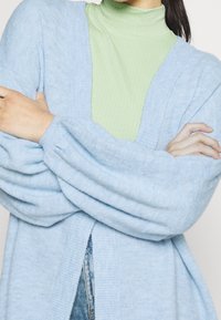 Světle modrý pletený kardigan s texturovanou tkaninou a širokými rukávy, navrstvený přes žebrovaný zelený rolák. Viditelné detaily šití.