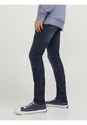 Persona con jeans ajustados azul oscuro, zapatillas de lona azul marino con cordones blancos y sudadera azul claro, de pie de lado.