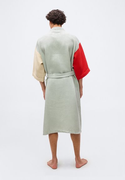 Άντρας όρθιος με γυμνά πόδια που φοράει ένα ρόμπα με υφή μήκους μέχρι το γόνατο, με μπεζ αριστερό μανίκι και κόκκινο δεξί μανίκι, φωτογραφημένος από πίσω.