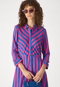 Robe chemise rayée rose et bleue avec des manches longues, un devant à boutons et un bustier ajusté. Accessoirisée avec des lunettes de soleil cat-eye transparentes et un sac à main bleu.