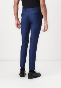 Pantalon bleu ajusté, en tissu lisse, avec deux poches arrière et des détails subtils. Associé à des chaussures en cuir noires.