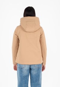 Tan softshell jack met een grote capuchon, een slank, aansluitend ontwerp en een gladde textuur, gedragen over casual blauwe jeans.
