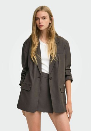 Ung kvinde med langt blondt hår iført en mørk oversized blazer og matchende nederdel over en hvid skjorte mod en ensfarvet baggrund.