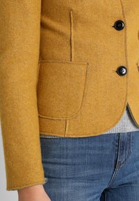Senfgelbe Wolle-Mix-Jacke mit schwarzen Knöpfen, zwei vorderen Taschen und abgerundetem Saum. Kombiniert mit grauem Pullover und Jeans.