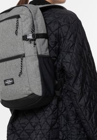 Person wearing black quilted jacket with gray Eastpak backpack featuring multiple zipped compartments and black straps.