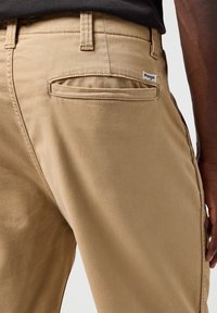 Beżowe bawełniane spodnie mają jedną tylną kieszeń, widoczne szwy i małą metkę Wrangler przy pasku. Tekstura wydaje się gładka.