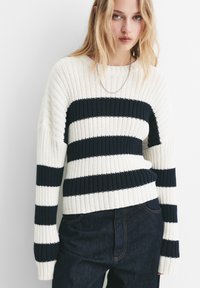 Jonge vrouw in een wit en zwart gestreept gebreid trui en een donkere spijkerbroek, staand tegen een witte achtergrond.