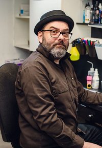 Muž s bradkou v hnědé košili na knoflíky sedí v černé židli, nosí černý klobouk a brýle, v pozadí jsou barevné pera.