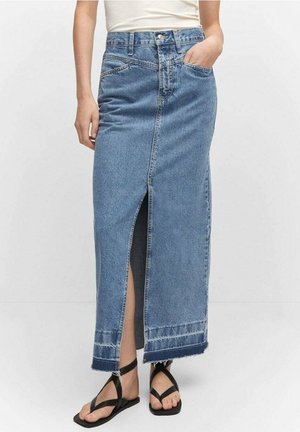 Kobieta ubrana w długą spódnicę jeansową o wysokim stanie w jasnoniebieskim kolorze, z rozcięciem z przodu i postrzępionym brzegiem, w połączeniu z czarnymi, płaskimi sandałami na paskach.