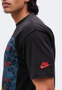 Νέος που φοράει μαύρο μπλουζάκι Nike με κόκκινο λογότυπο στο μανίκι και γραφικό σχέδιο μπροστά, κοιτάζοντας προς τα αριστερά με ορατή γνάθο και ώμο.