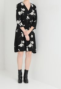 Vestido negro envolvente con estampado floral blanco, escote en V, mangas tres cuartos y cinturón anudado en la cintura; combinado con botines negros.
