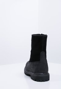 Schwarze Stiefeletten aus Wildleder mit Gummisohle; verfügen über eine glatte Oberfläche und einen niedrigen Absatz. Die Rückansicht zeigt ein Branding-Detail.