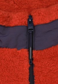 Rode fleecestof met een gestructureerd uiterlijk, in contrast met een gladde marineblauwe stofpanelen. Voorzien van een zwarte rits en trekkoorddetails.